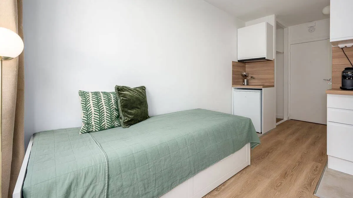 Lit avec couvre-lit vert et kitchenette en arrière-plan